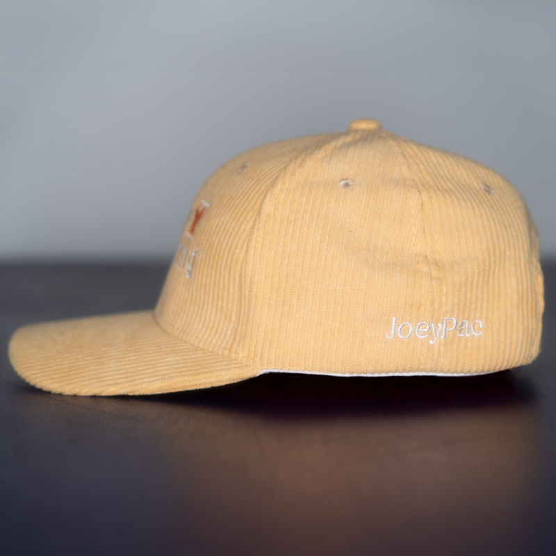 The side of a yellow beige corduroy baseball cap with 'JoeyPac' embroidered in white on the lower portion of the hat.